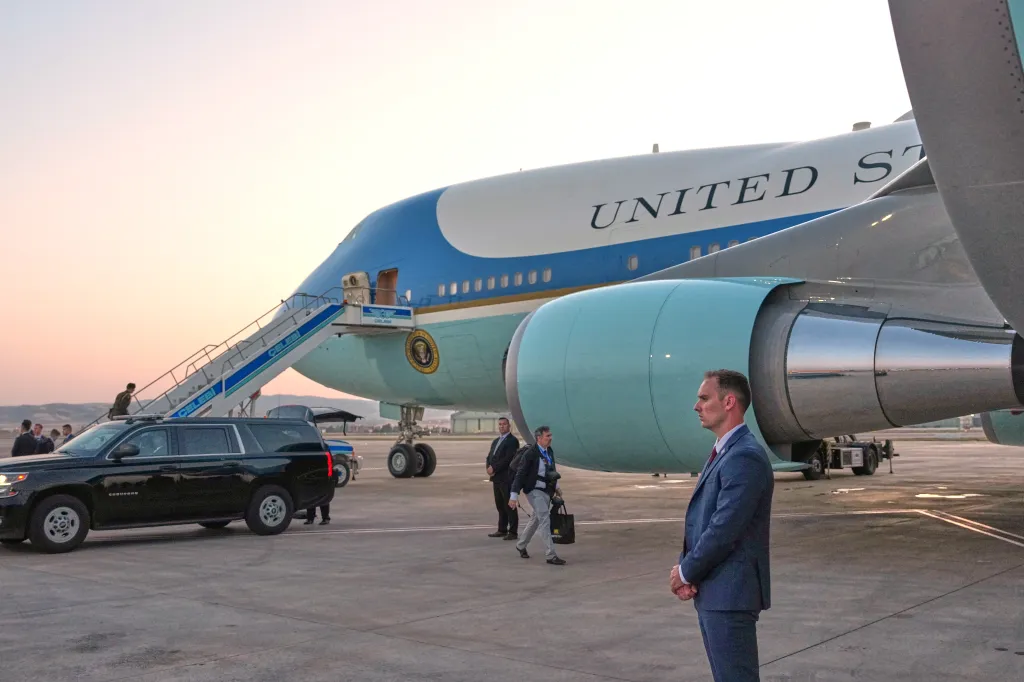 Air Force One parked on the tarmac with a catering truck, a black SUV, and several people nearby, including a man in a suit in the foreground.