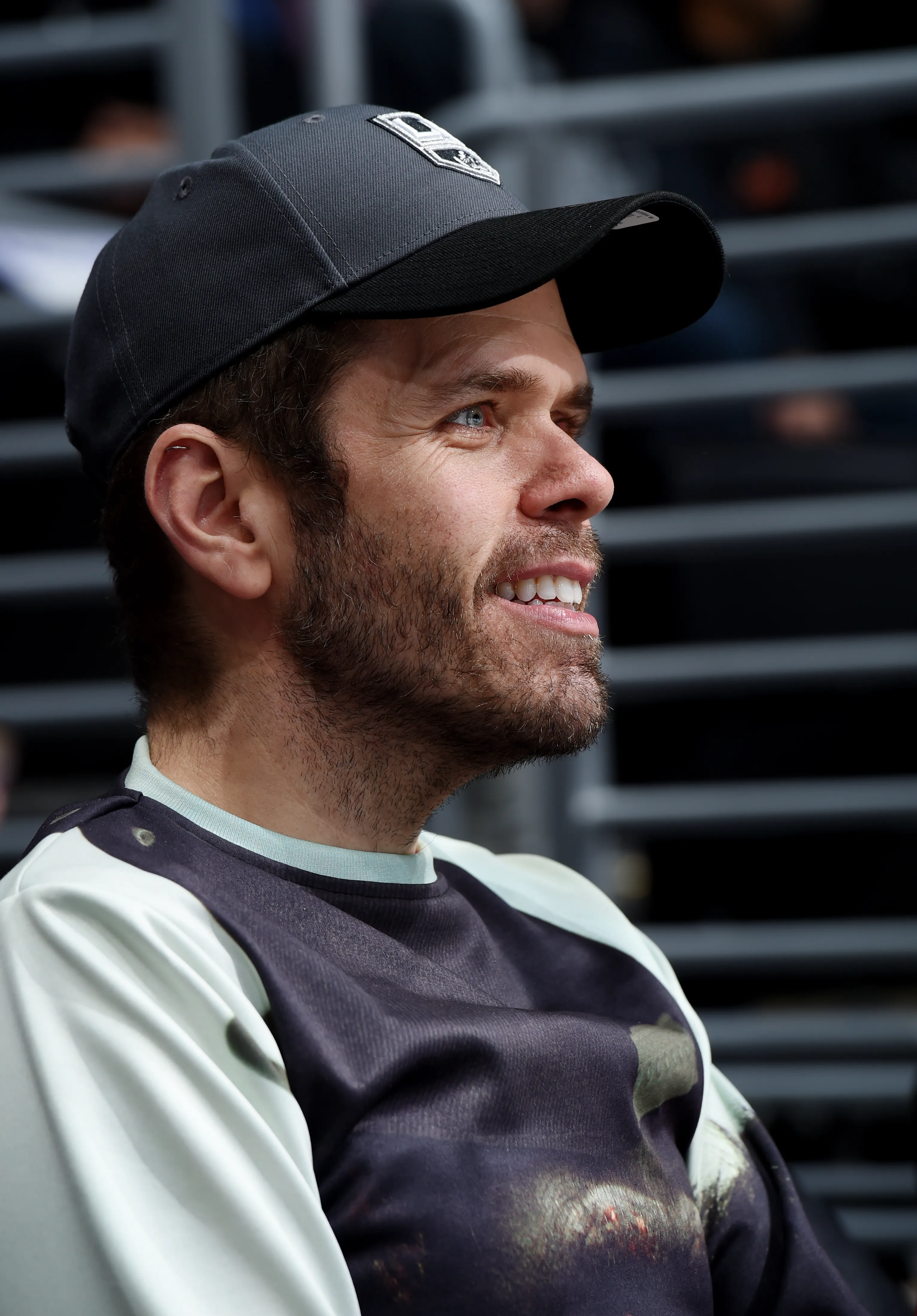 Perez Hilton in a black baseball cap and a patterned shirt, watching a game.