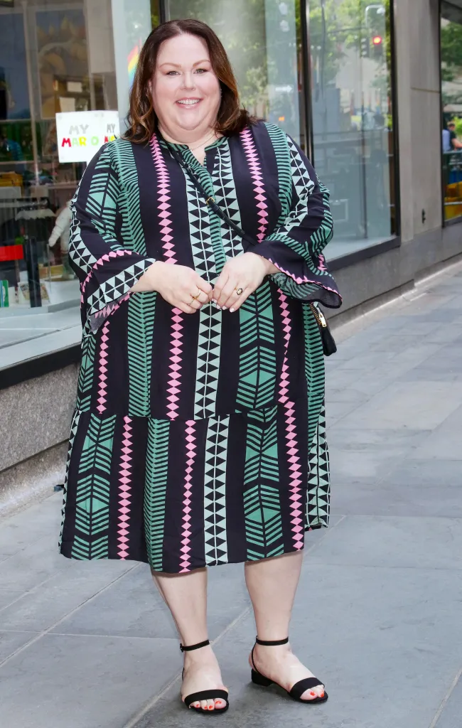 Chrissy Metz smiling while posing in a black, pink, and teal patterned dress and black sandals.