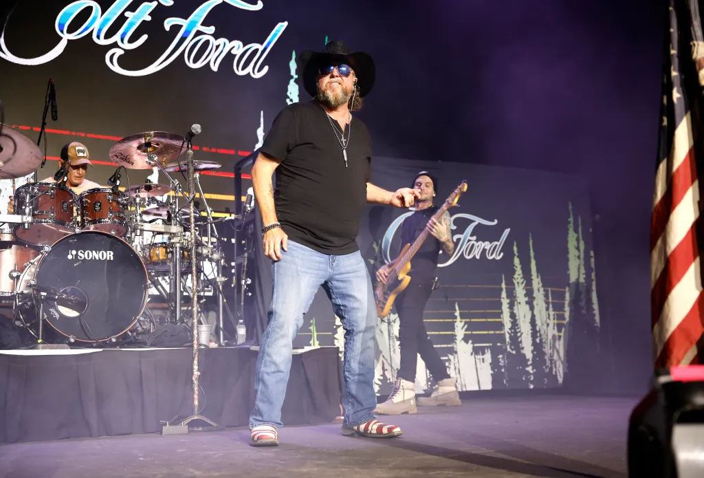 Colt Ford performing on stage with a drummer, bassist, and American flag nearby.