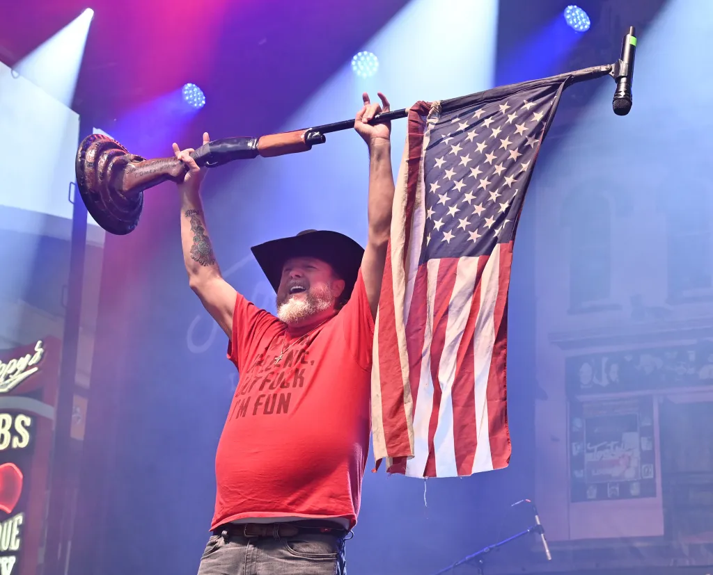 Colt Ford holding a microphone stand with an American flag attached to it above his head on stage.