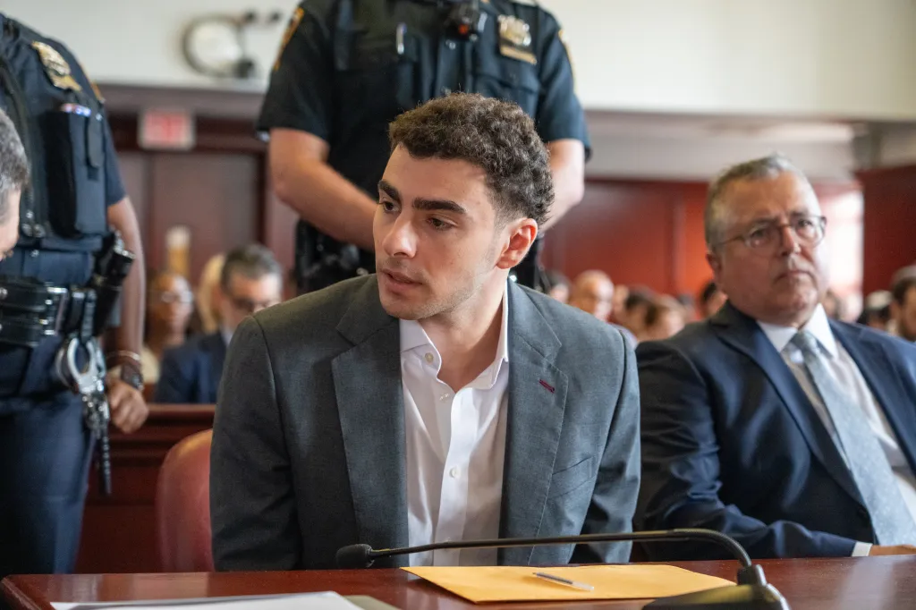 Luigi Mangione appearing in a New York City court, with a police officer standing behind him.