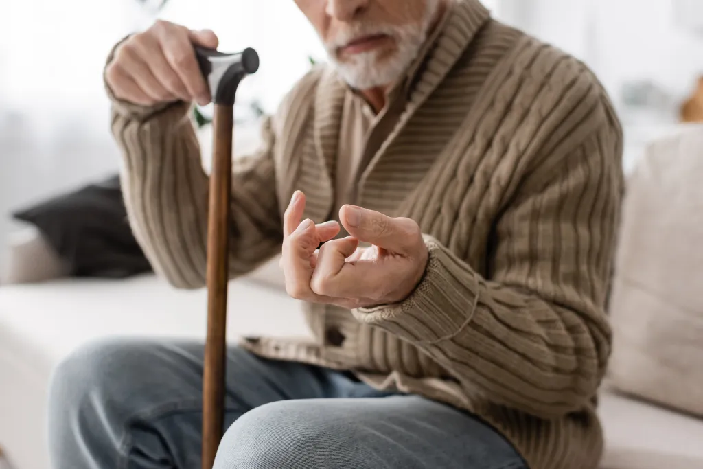 Cropped view of a senior man with Parkinson's disease holding a walking cane while sitting on a couch at home.