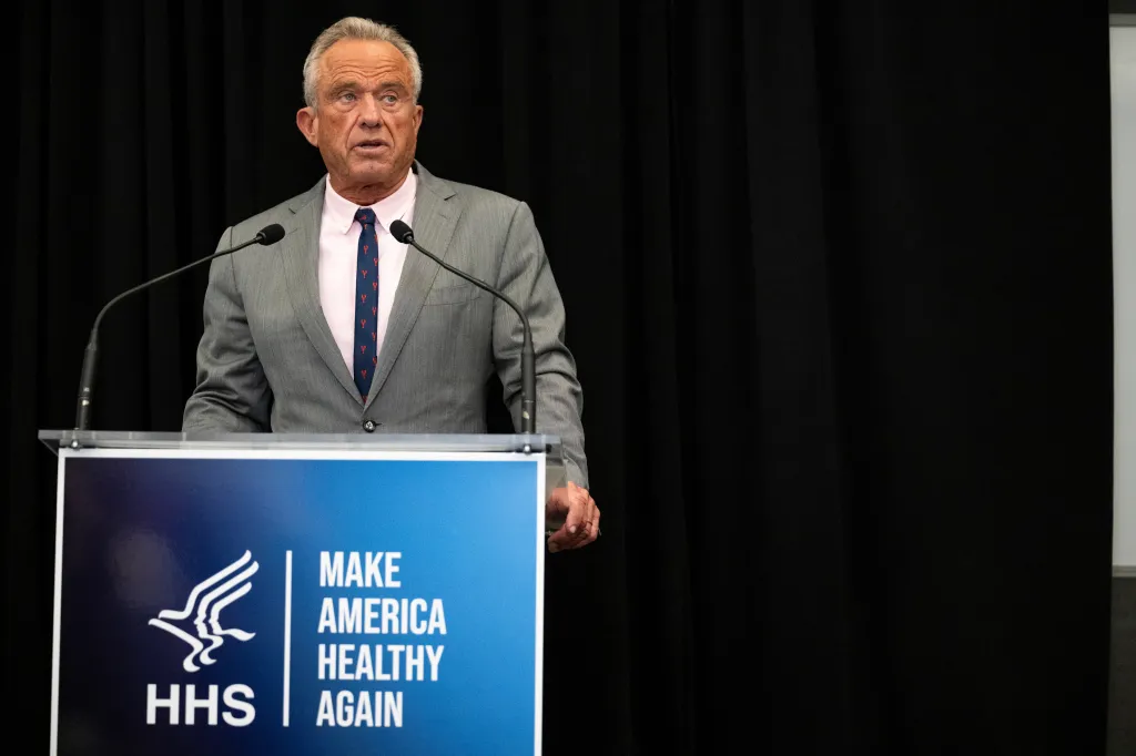 Robert F. Kennedy Jr. speaking at a podium with the HHS logo and "Make America Healthy Again" slogan.