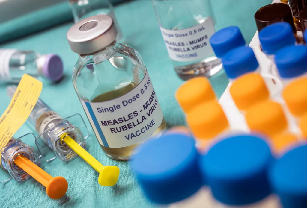 Vials labeled "MEASLES - MUMPS RUBELLA VIRUS VACCINE" and syringes on a blue surface.