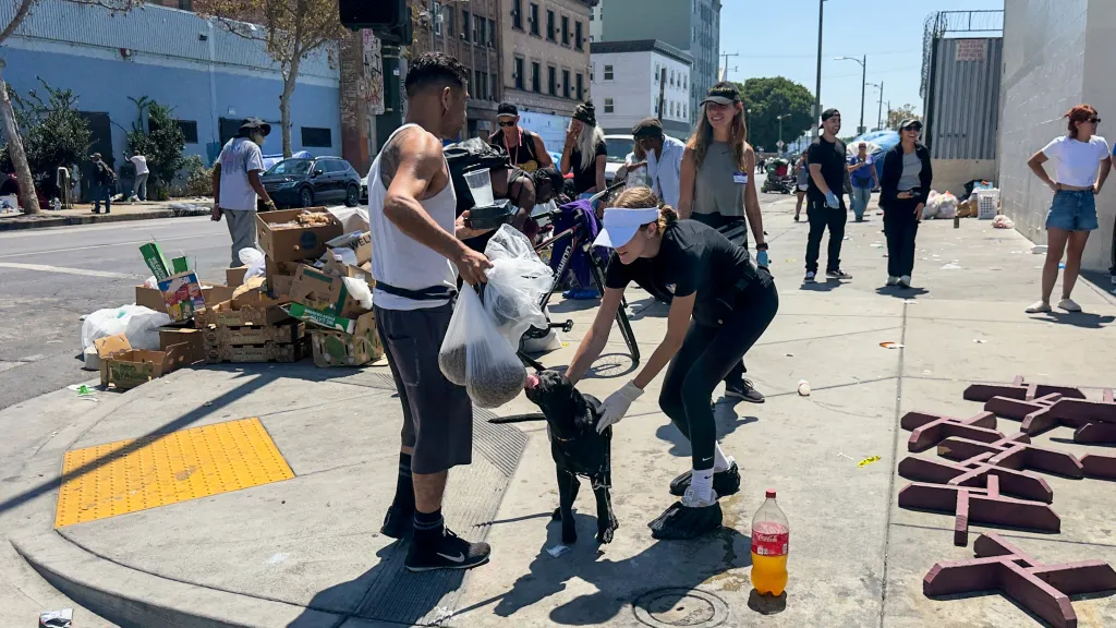 A woman in a visor and gloves feeds a black dog while a man holding plastic bags of dog food watches, with several people and cardboard boxes visible on a dirty street.