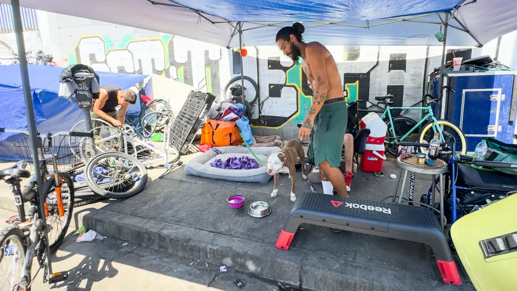 A man and his dog stand under a blue canopy on a sidewalk filled with various belongings, while another man works on a bicycle nearby.