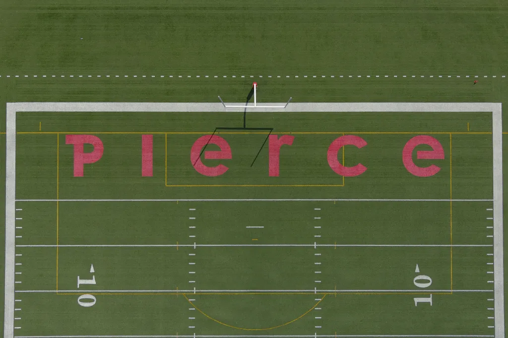 Aerial view of the "PIERCE" painted in red on the end zone of a green football field at John Shepard Stadium, with goalposts and yard lines visible.