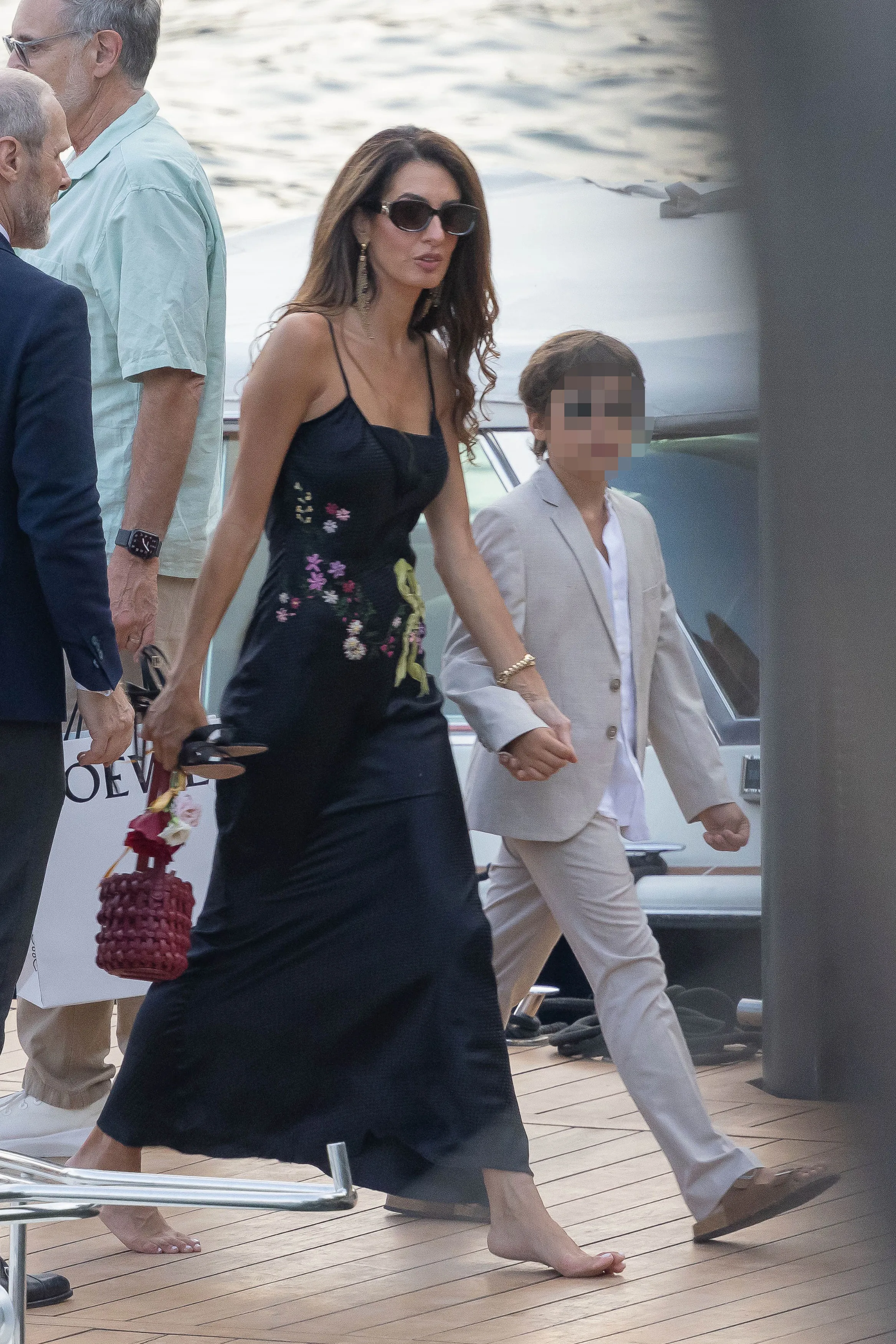 Amal Clooney walking barefoot in a black floral dress, holding hands with Alexander Clooney in a light suit.