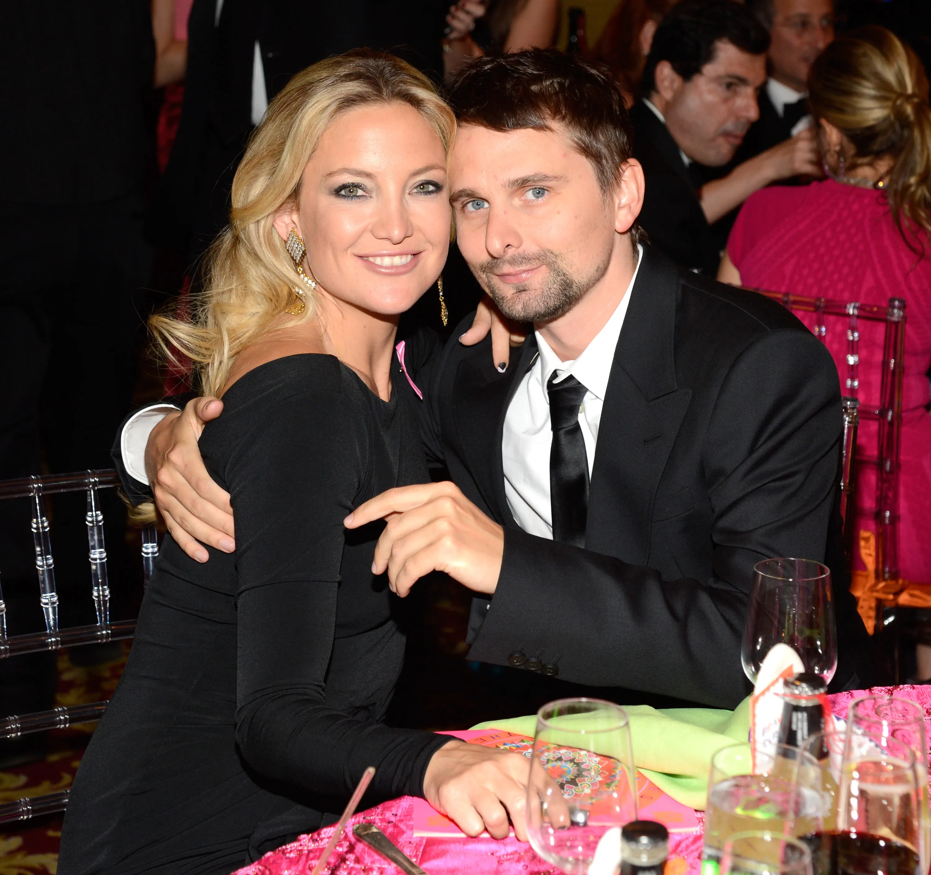 Kate Hudson and Matt Bellamy pose for a photo, with Hudson wearing a black dress with a pink ribbon pin and Bellamy in a black suit.