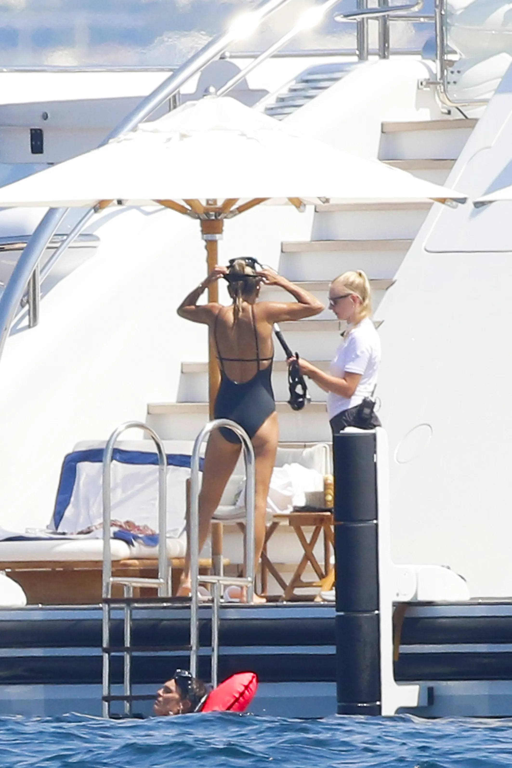 A woman in a black swimsuit adjusts her snorkeling mask on a yacht while another person holds equipment and Tom Brady is in the water with a red buoy.