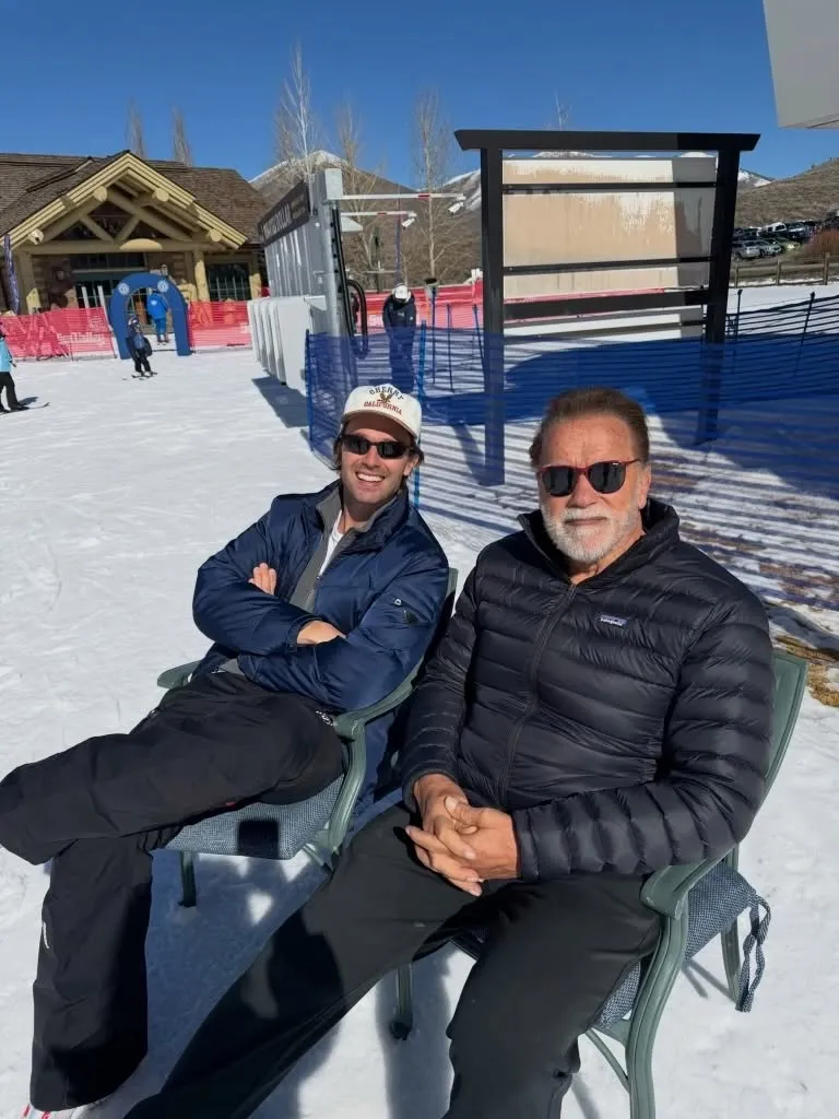 Arnold Schwarzenegger and his son Patrick Schwarzenegger smile for a photo while sitting in chairs on a snowy day.