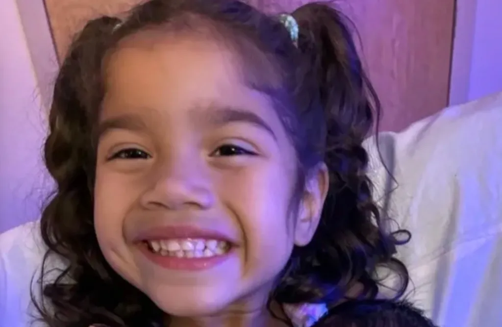 A young girl, Lailah, with dark curly hair in pigtails, smiles broadly.