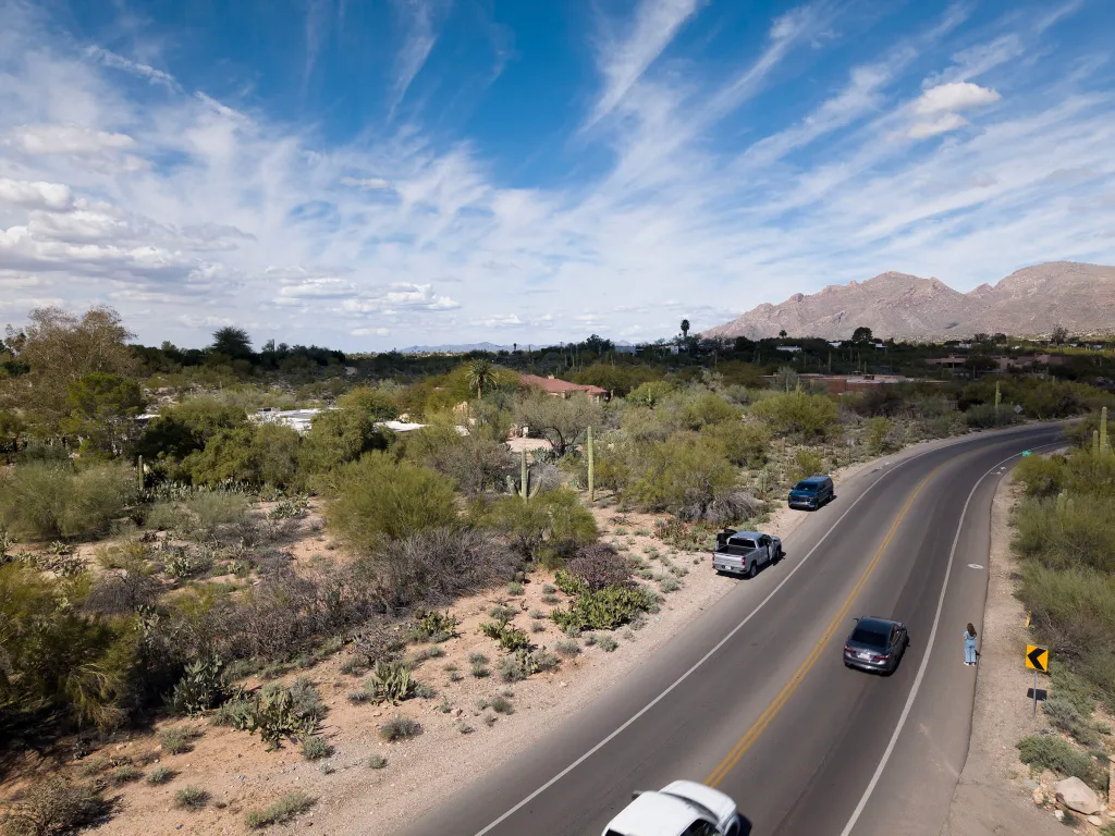 Overhead view of a desert road with mountains in the background, a woman in jeans on the side, and cars driving past.