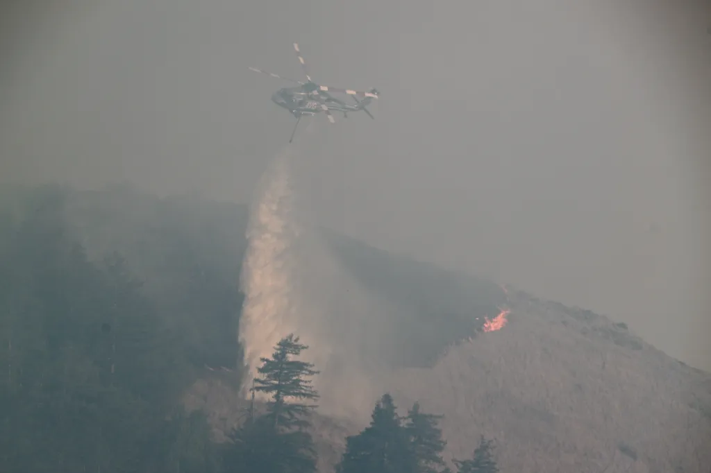 A helicopter drops water on a wildfire in Big Sur, California, with visible flames on a distant hillside.