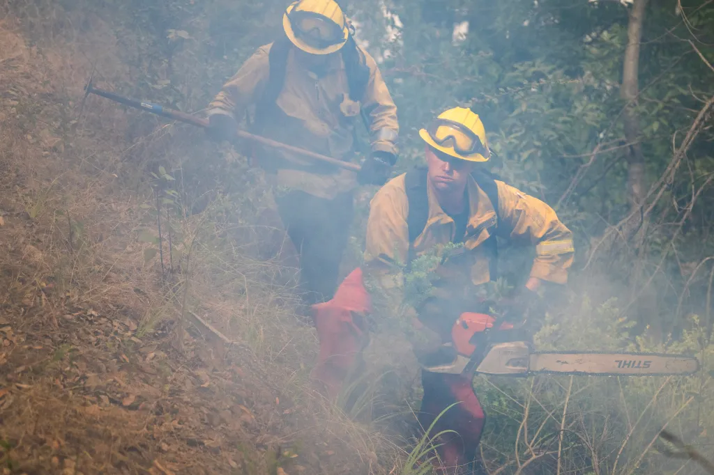 A firefighter uses a chainsaw to cut brush while smoke fills the air.