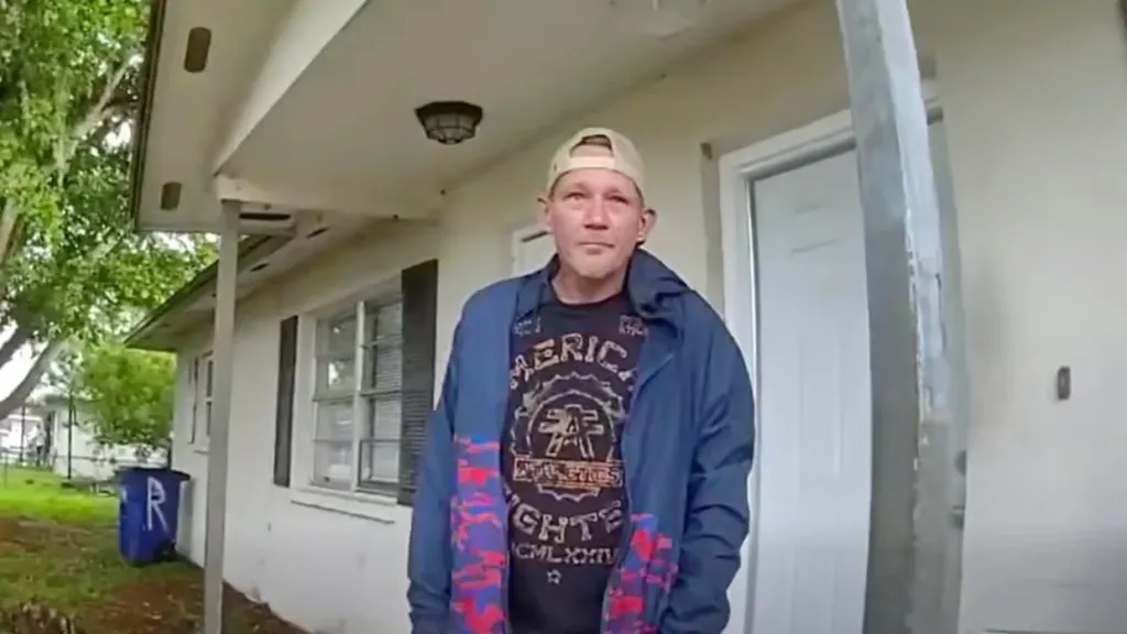 A man in a baseball cap, blue jacket, and "American Fighters" t-shirt stands on a porch.