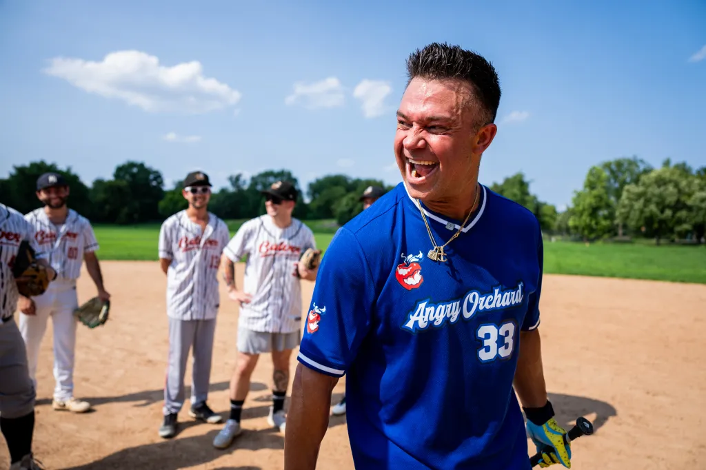 Baseball legend Nick Swisher in an Angry Orchard jersey smiles on a baseball field with other players in the background.