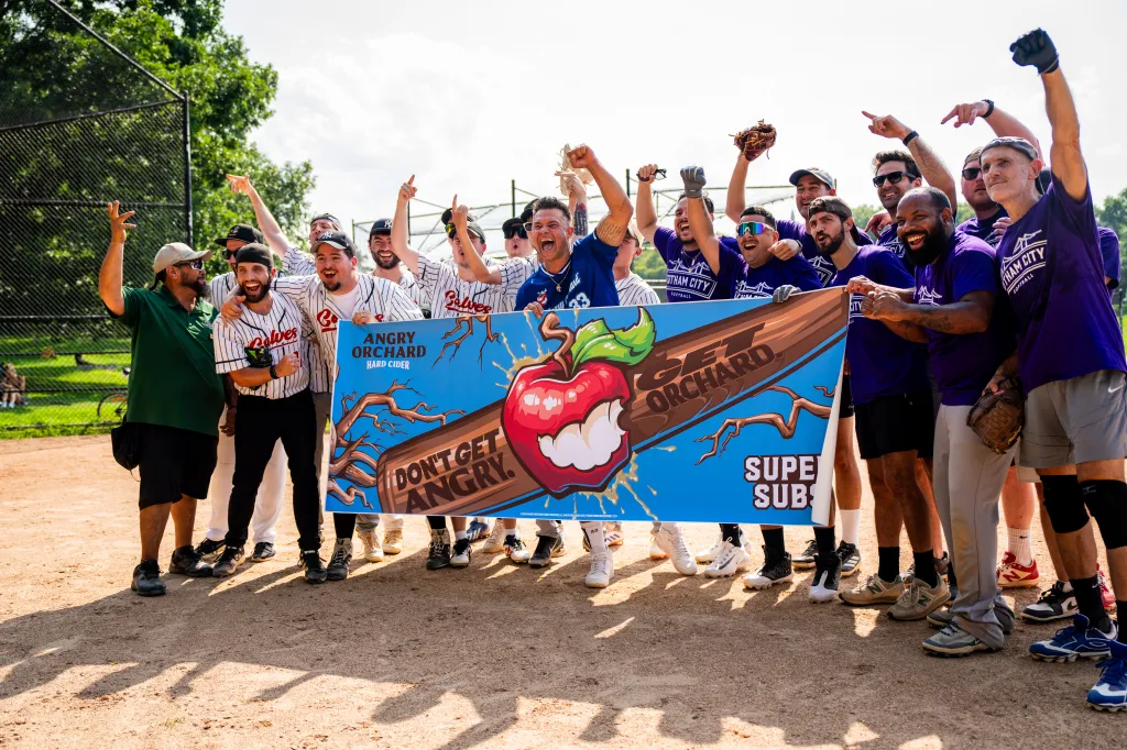 Nick Swisher and the Sons of Pitches softball team cheer while holding an Angry Orchard Super Subs banner on a baseball field.