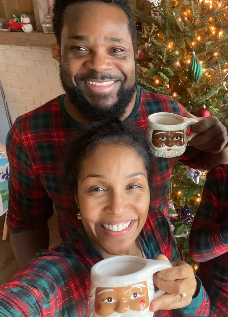 Smiling man and woman in plaid pajamas holding Santa mugs by a Christmas tree.