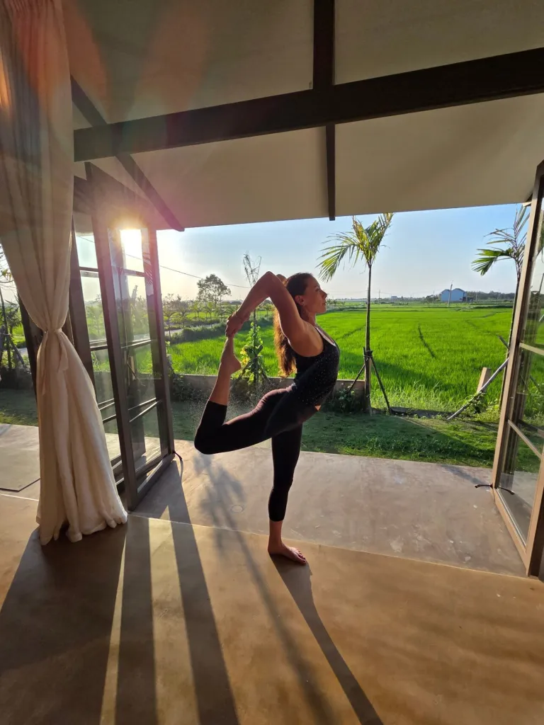 Chaz in a black top and pants holding a yoga pose on a patio overlooking a rice field.