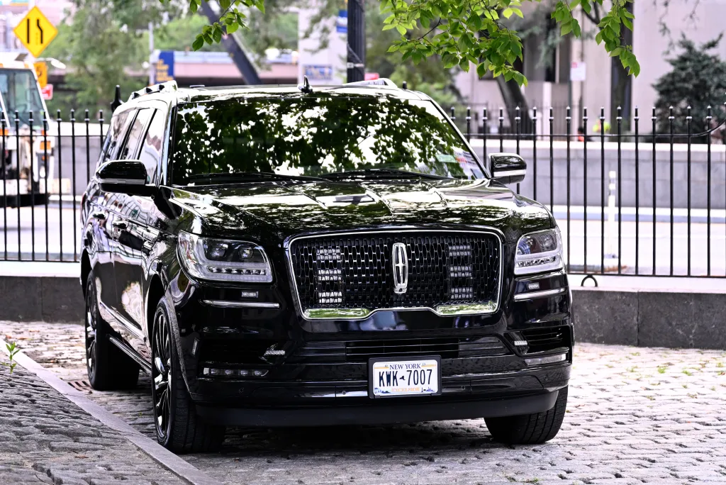 A black Lincoln Navigator with a New York license plate "KWK-7007" parked on cobblestones in front of City Hall.