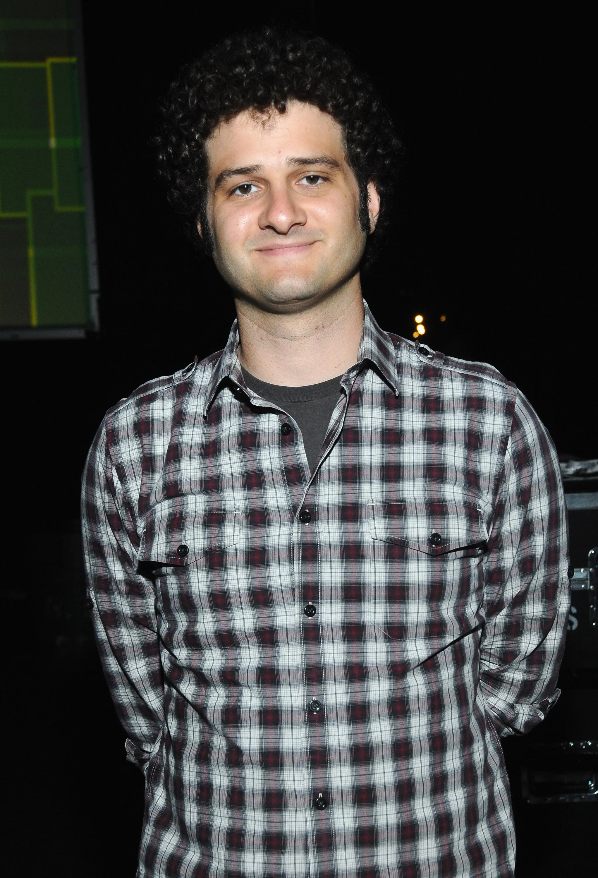 a man with curly brown hair and a black plaid shirt stands in front of a black background.