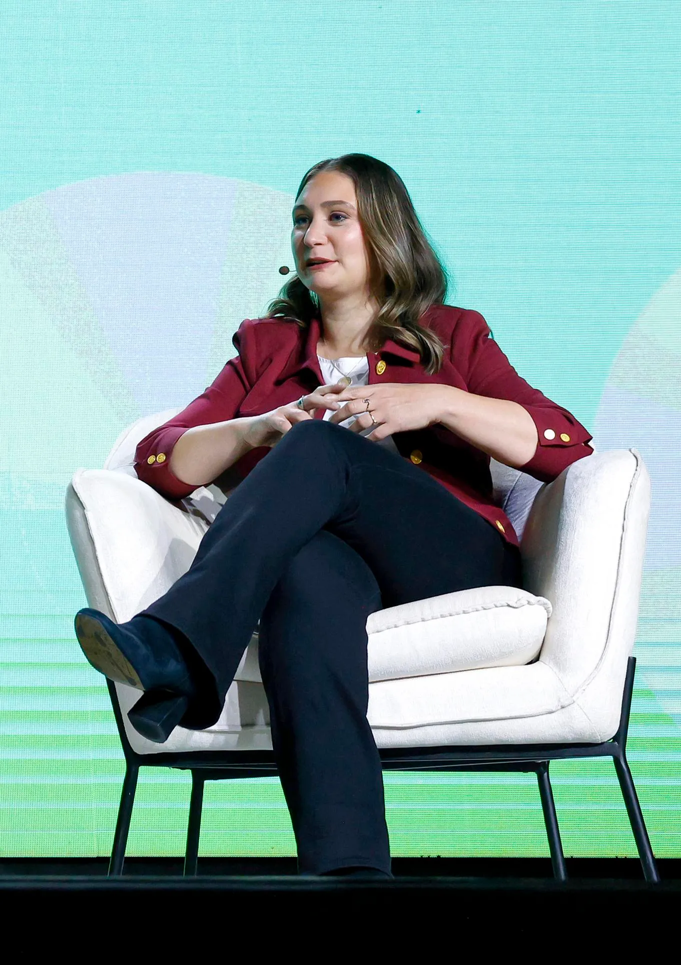 a woman with long brown hair, wearing a red jacket, dark. blue jeans, and black shoes sits in front of a blue-green screen in the background.