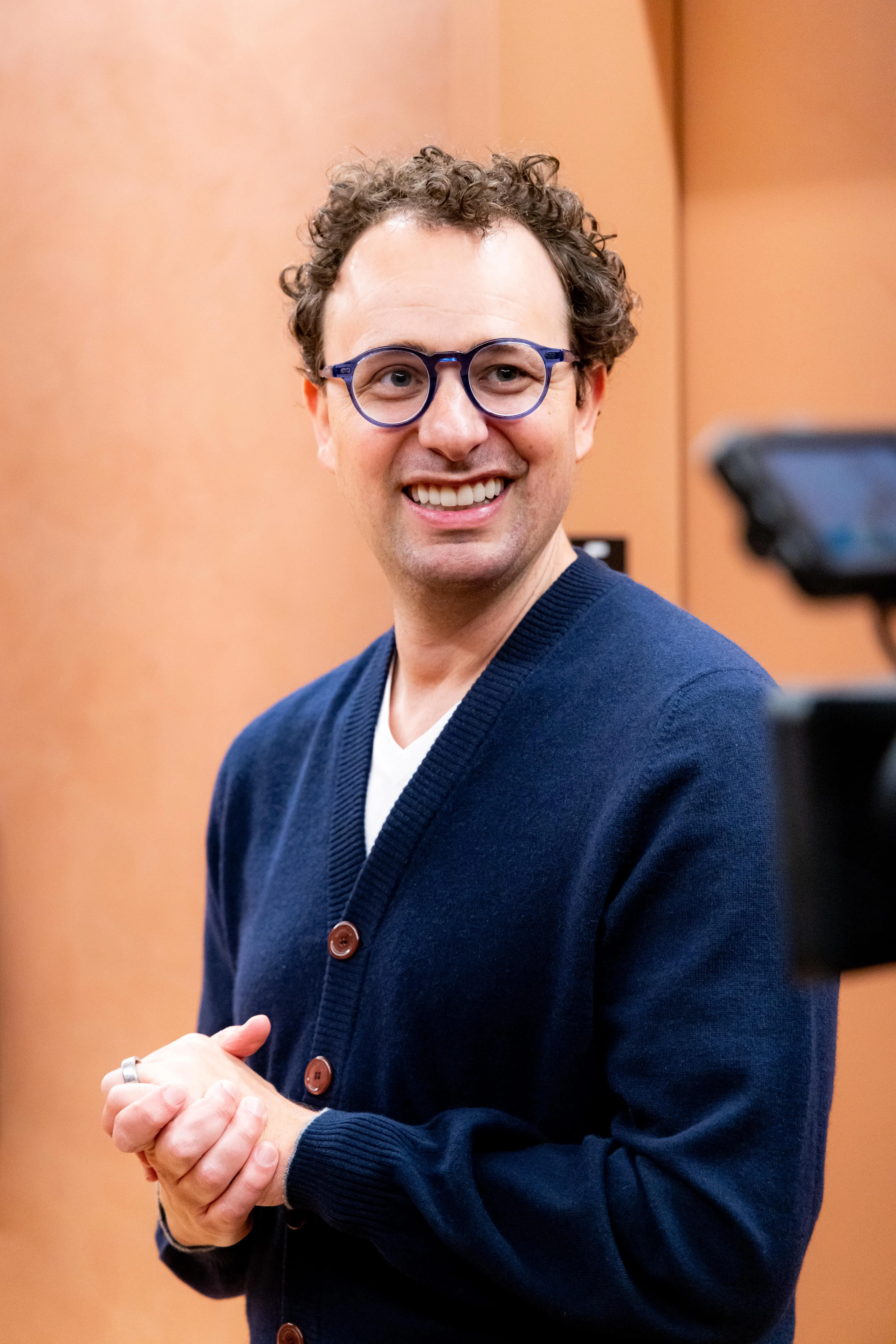 a man with curly brown hair and blue glasses, wearing ab lue sweater, smiles and stands in front of an orange wall.