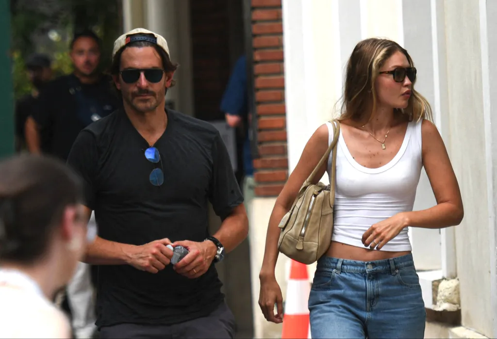 Gigi Hadid in a white top and blue jeans next to Bradley Cooper in a black t-shirt and backward cap, both wearing sunglasses.