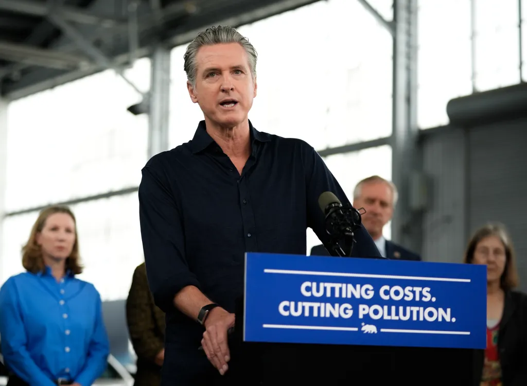 Governor Gavin Newsom speaking at a press conference in Oakland, California, with a sign that reads "Cutting Costs. Cutting Pollution."