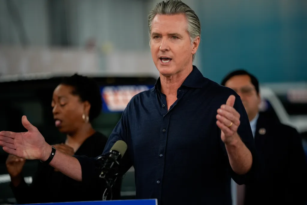 Governor Gavin Newsom speaking at a press conference in Oakland, California, with a microphone in front of him and two blurred figures behind him.