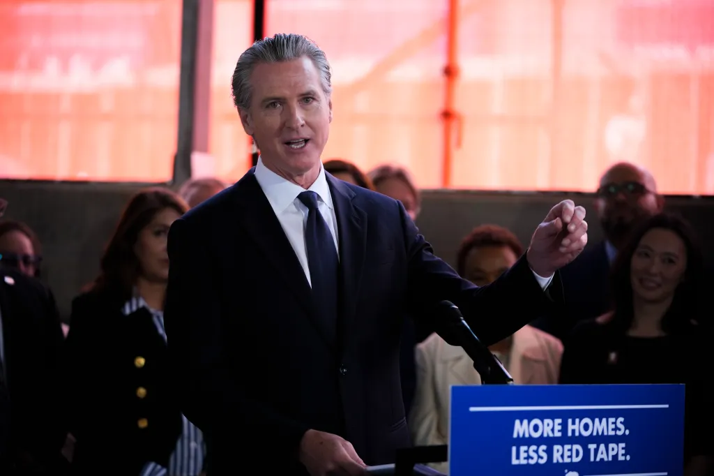 Governor Gavin Newsom speaking at a press conference in Oakland with a sign in the foreground that reads "MORE HOMES. LESS RED TAPE."