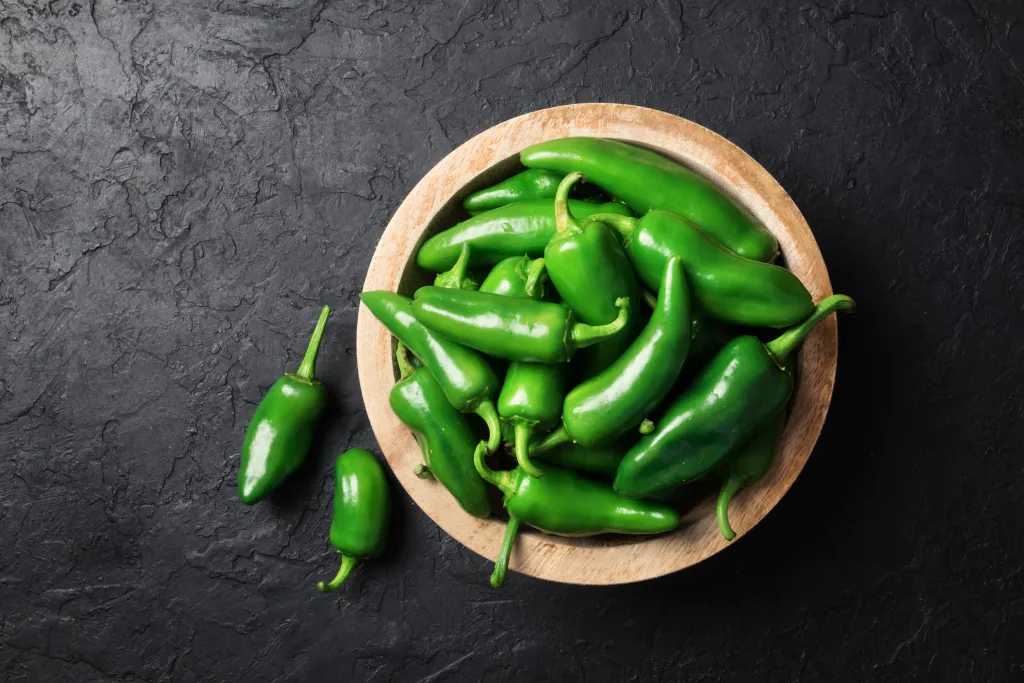 A wooden bowl filled with green jalapeno peppers, with two peppers outside the bowl, on a dark textured surface.