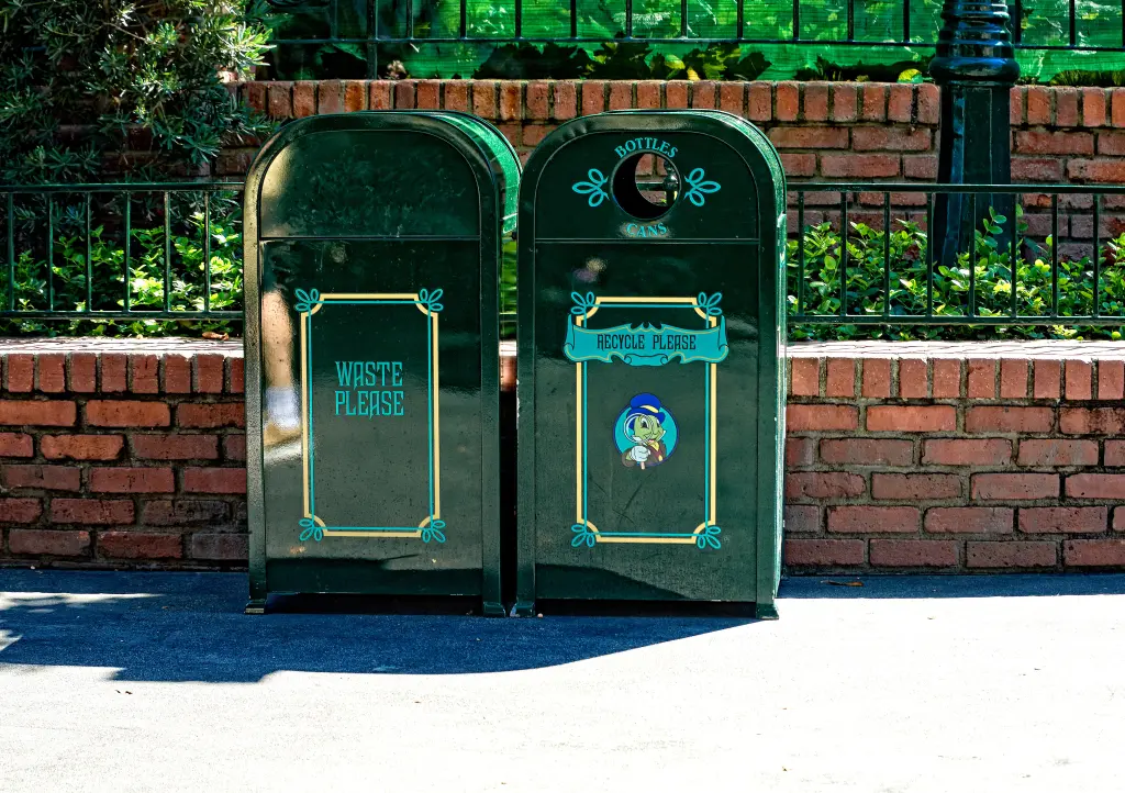 Two dark green recycling bins, one for "waste" and one for "recycle," stand on a paved path in a Universal Studios Theme Park.