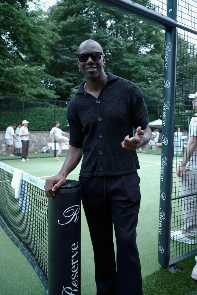 Terrell Owens smiling, wearing sunglasses, and leaning on a tennis net post.