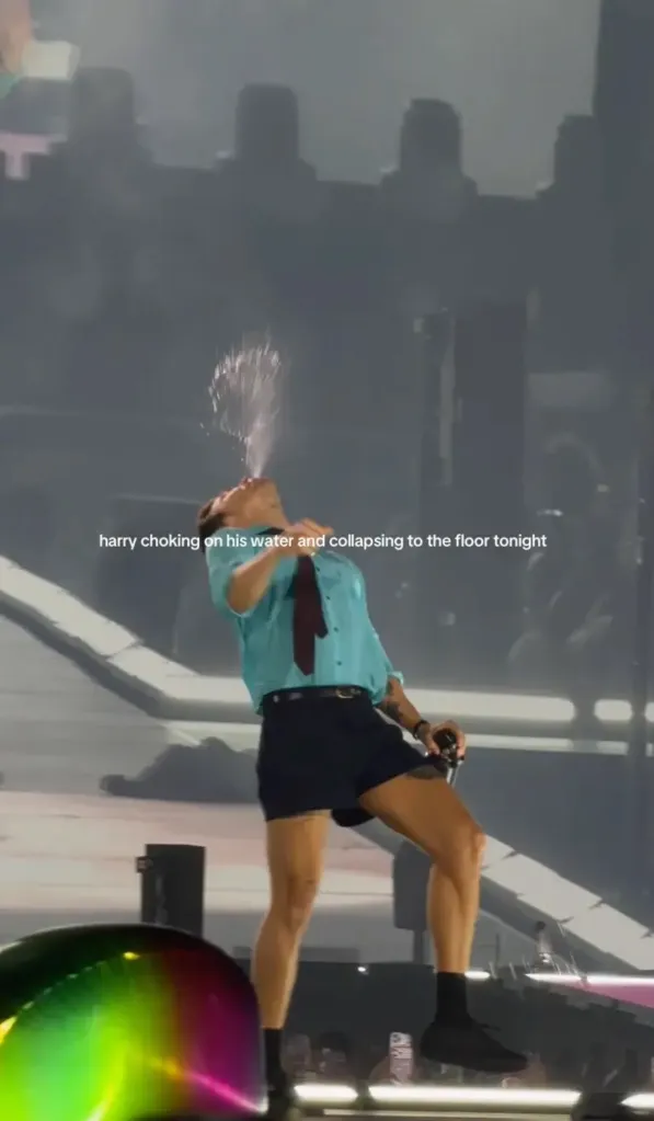 Harry Styles choking on water, wearing a blue shirt, black shorts, and a dark tie on stage at a concert.