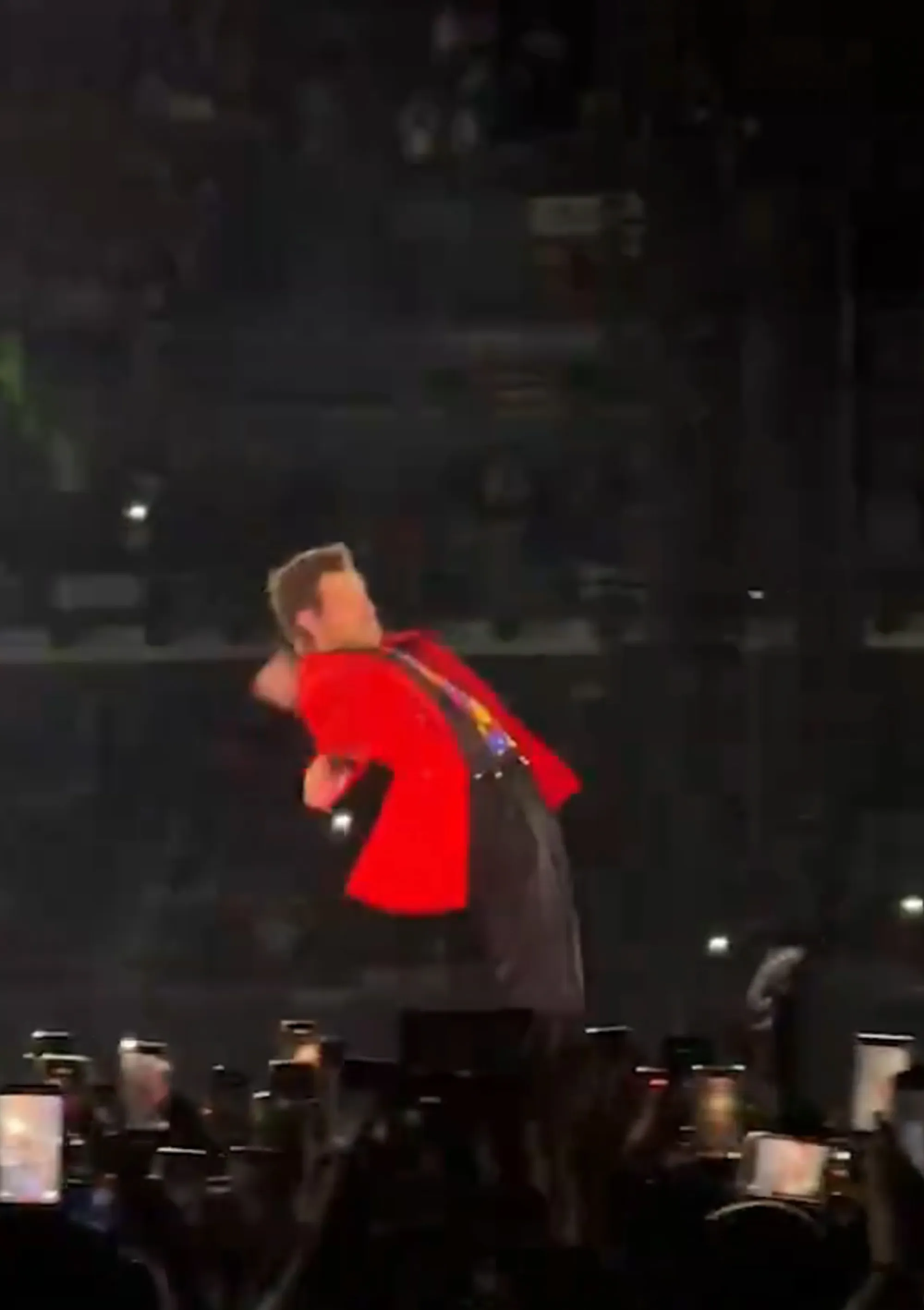 Harry Styles on stage, wearing a red blazer, black pants and a colorful shirt, leans back almost falling while performing in front of a crowd.