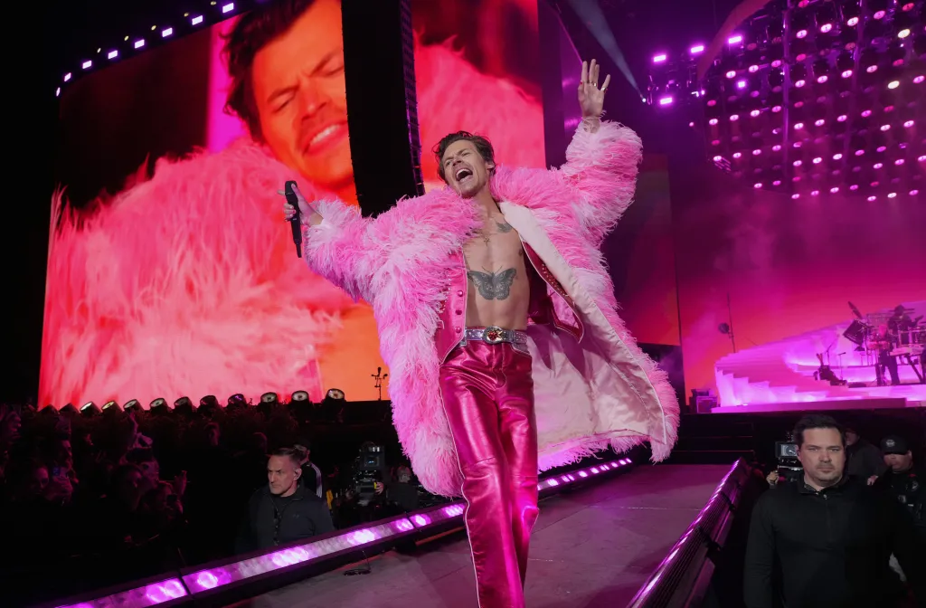 Harry Styles performing on the Coachella stage in a pink feathered coat, shiny pink pants, and a butterfly tattoo on his chest, with a large screen behind him showing a close-up of his face.