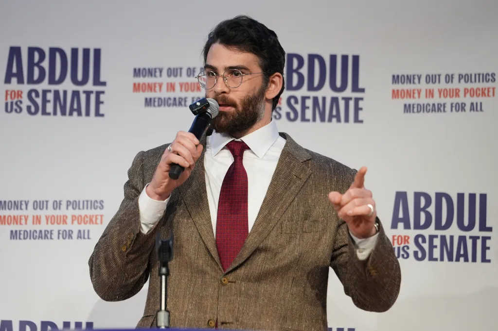 Hasan Piker speaking at a campaign rally for Abdul El-Sayed, with "Abdul for US Senate" and campaign slogans visible in the background.