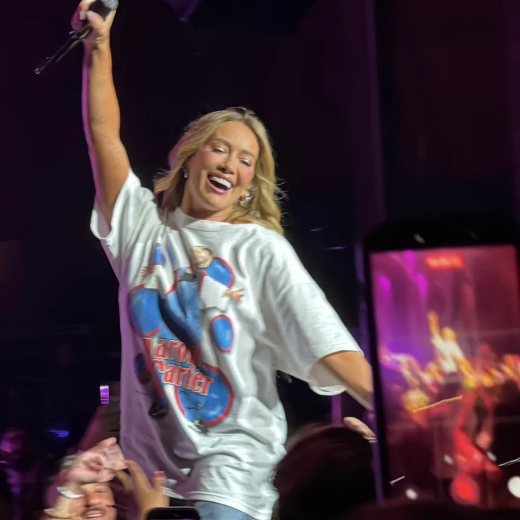 Hilary Duff performing on stage, wearing a white Aaron Carter tribute t-shirt.