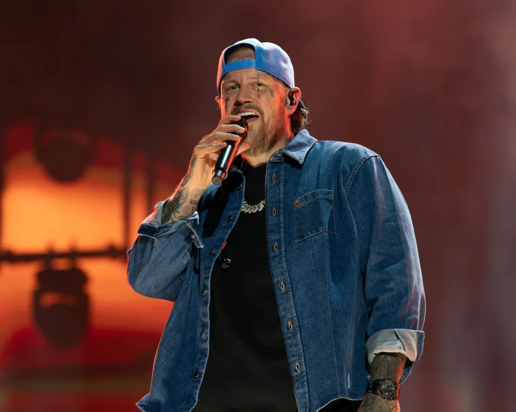 Jelly Roll performing on stage, wearing a blue cap and denim shirt, holding a microphone.