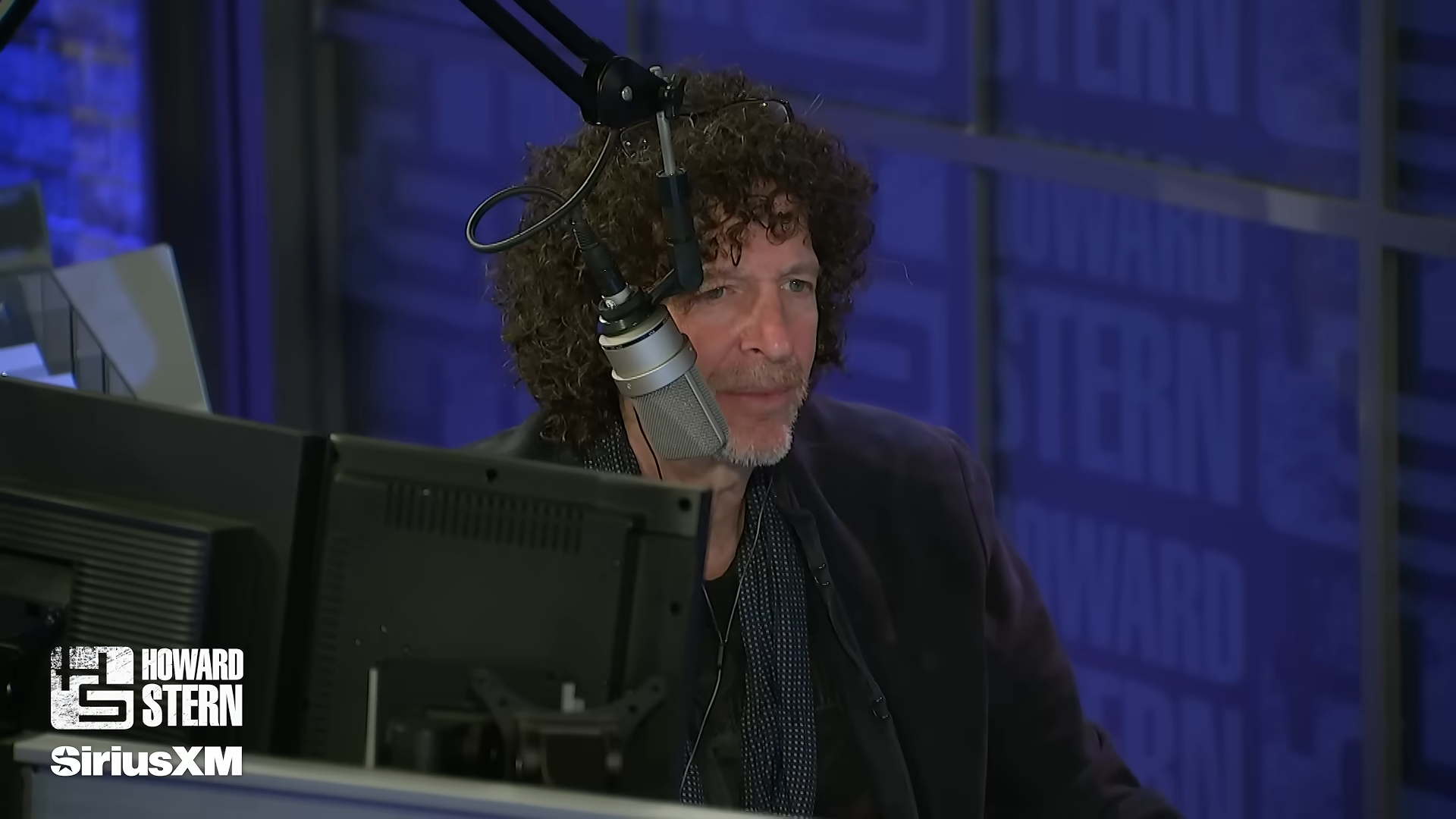 Howard Stern at a microphone, with a Howard Stern SiriusXM logo in the bottom left corner.
