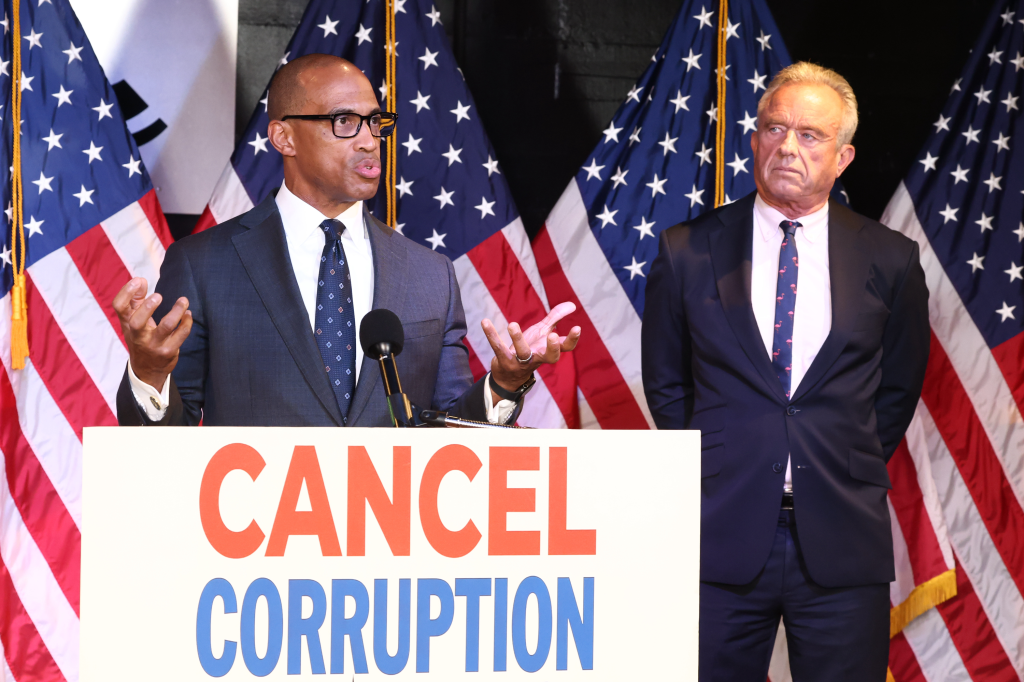 HUD Secretary Scott Turner speaking at a podium next to HHS Secretary Robert F. Kennedy, Jr. with "CANCEL CORRUPTION" visible on the podium.