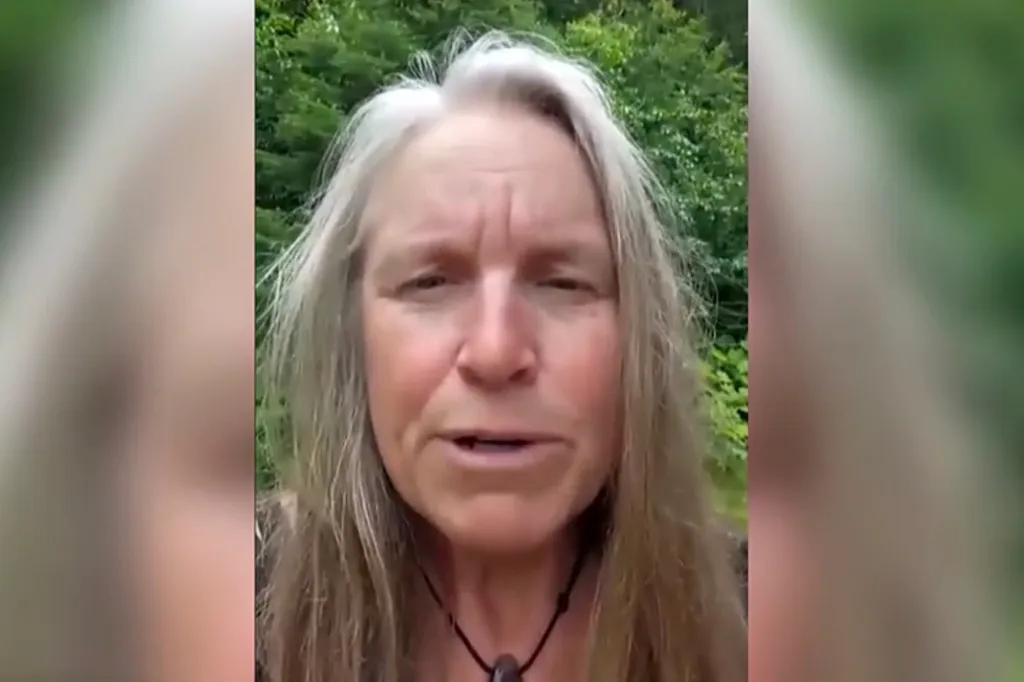 A woman with long gray hair and a necklace with a dark stone pendant speaks into the camera, with green foliage in the background.