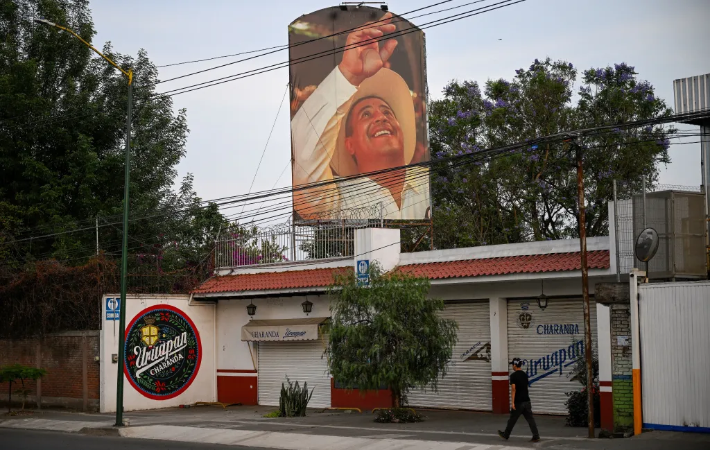 A man walks past a large portrait of assassinated Municipal President of Uruapan Carlos Manzo on a building, with closed storefronts and power lines in the foreground.