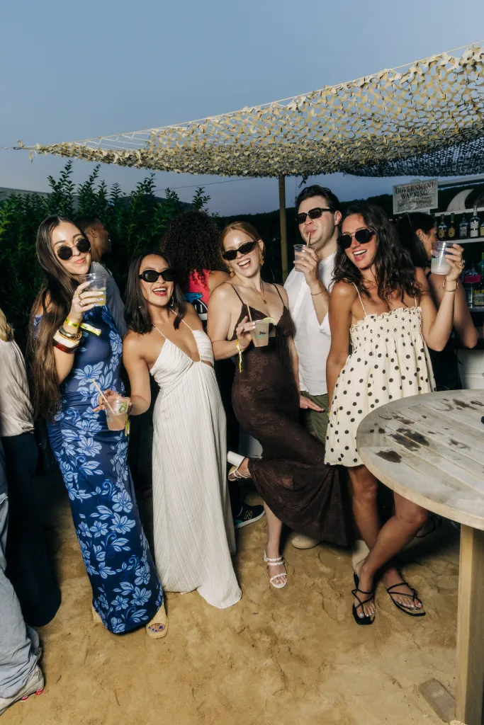 Five people wearing sunglasses and holding drinks pose on a sandy ground under a camouflage net at an outdoor party.
