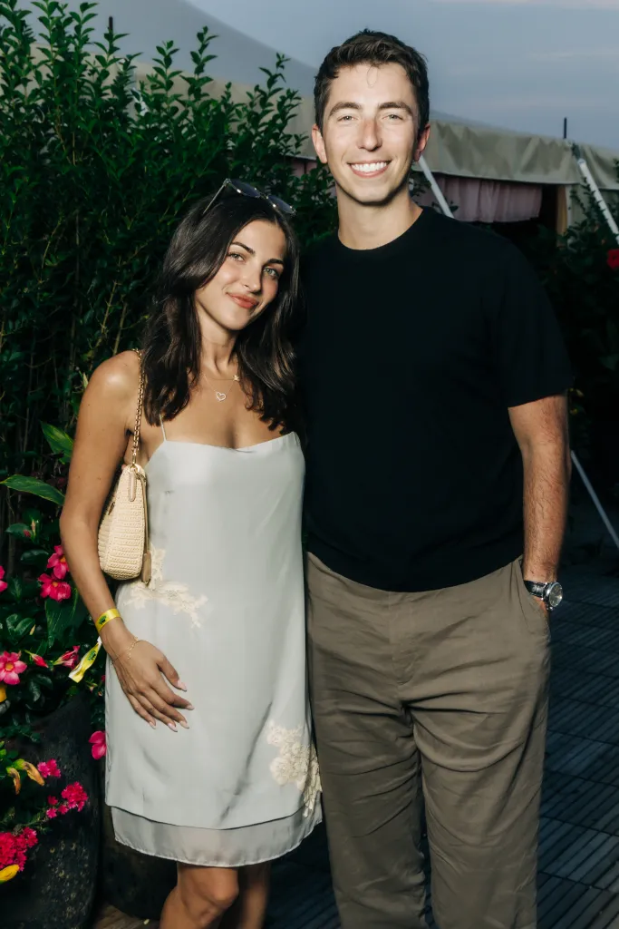 A man and a woman stand smiling, the woman in a light blue dress with a woven bag, and the man in a black shirt and khaki pants.
