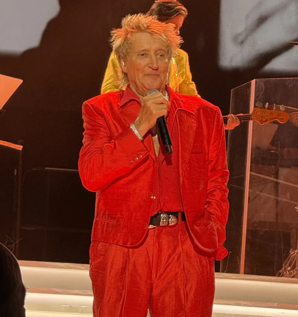 Rod Stewart performing live in New York, wearing a red suit and holding a microphone.
