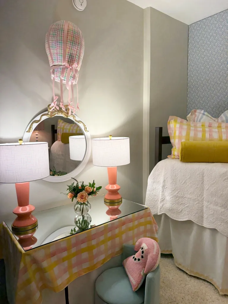 A dorm room decorated with a white bed, pink and yellow pillows, a vanity with pink lamps, and a checkered tablecloth.